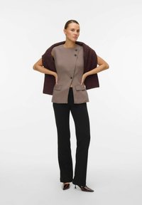 Blazer senza maniche marrone su misura con due tasche frontali e pantaloni neri. Il modello drappeggia un scialle bordeaux sulle spalle, in piedi su uno sfondo neutro.