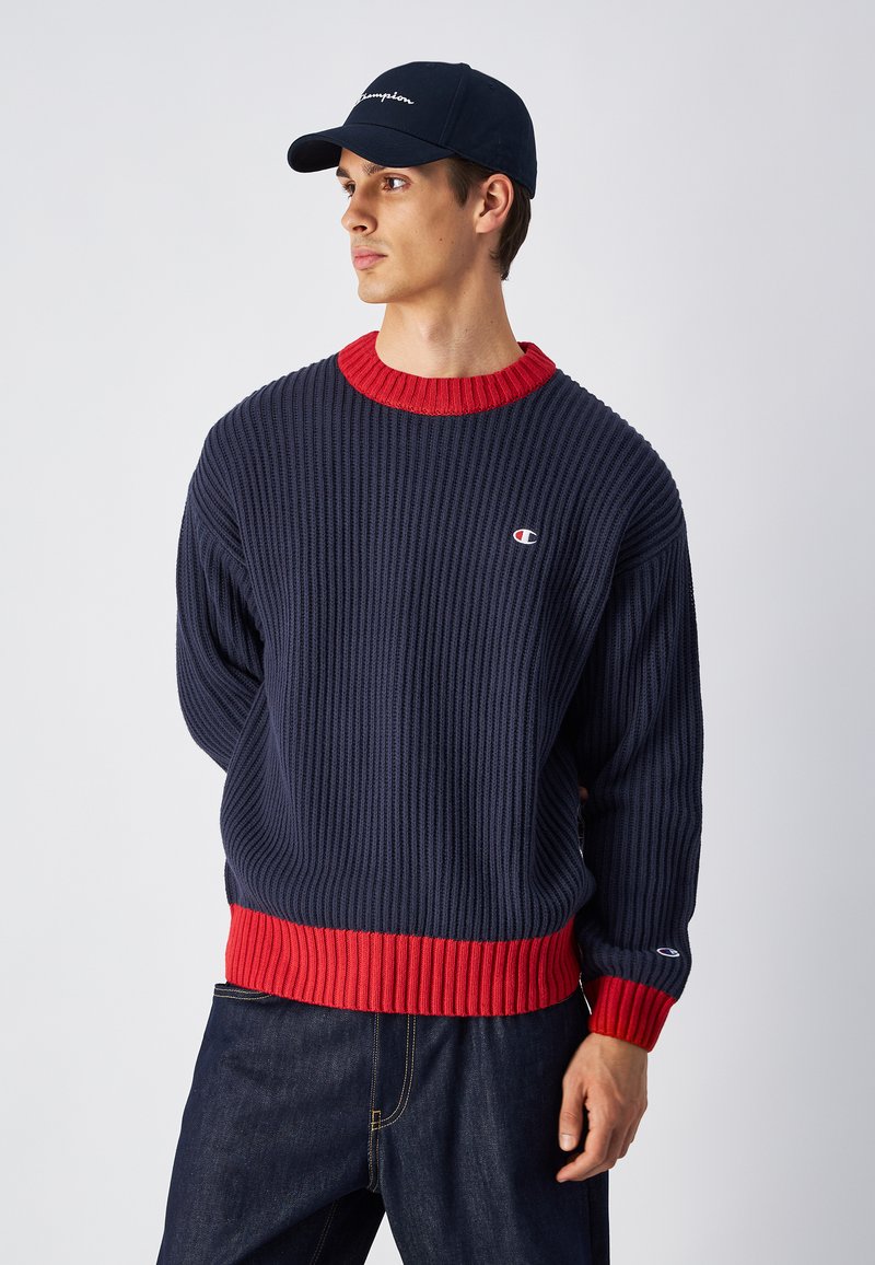 Pull en maille côtelée marine avec des accents rouges au niveau de l'encolure et de l'ourlet, arborant un petit logo sur la poitrine gauche. Associé à un pantalon en denim foncé.
