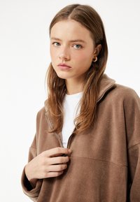 Brun fleece pullover med høj krave og halvlynlås, der har en blød tekstur og en afslappet pasform. Bæres over en hvid skjorte.