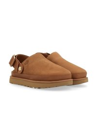 Zapatos slip-on de ante marrón con una suela de goma gruesa, punta redonda y correa trasera ajustable con un acento de botón de metal.