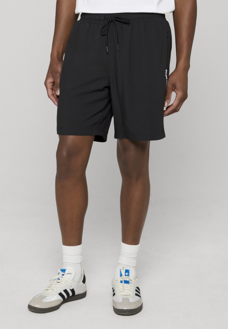 Personne portant un short noir à cordon, des chaussettes blanches montantes et des baskets blanches à rayures noires, debout sur une surface gris clair.