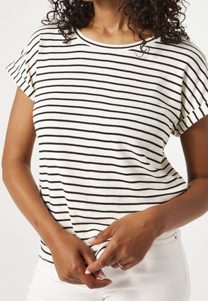 Nainen, jolla on kiharat hiukset, valkoinen lyhythihainen paita, jossa mustat vaakaraitoja, ja valkoiset housut, kädet rentosti edessä yhteen kiedottuina.