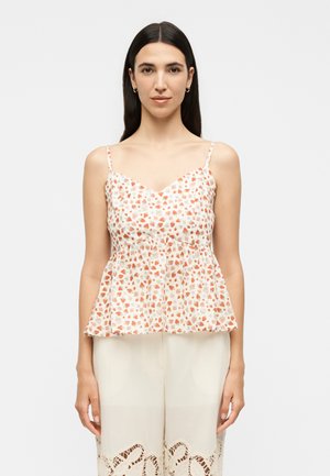 Kobieta o długich ciemnych włosach nosi biały top bez rękawów z małym pomarańczowo-zielonym kwiatowym wzorem oraz kremowe spodnie z koronkową lamówką.