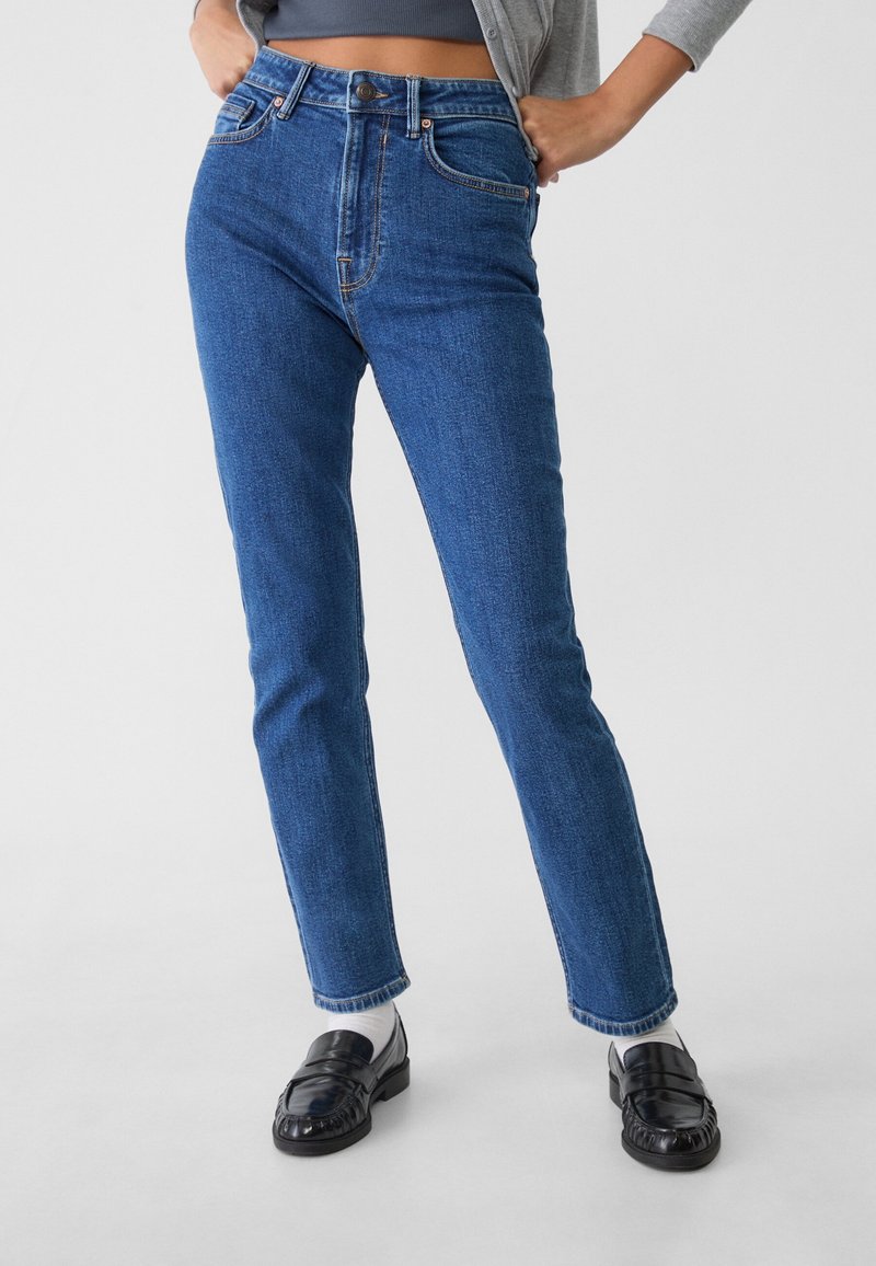 Femme portant un jean bleu taille haute à jambes droites, chaussettes blanches, mocassins noirs, cardigan gris et top court foncé, debout les mains sur les hanches.