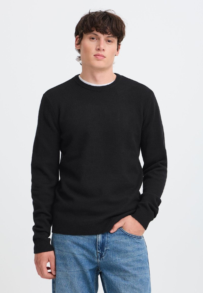 Giovane uomo con capelli scuri che indossa un maglione nero a maniche lunghe e jeans blu, in piedi davanti a uno sfondo bianco semplice.