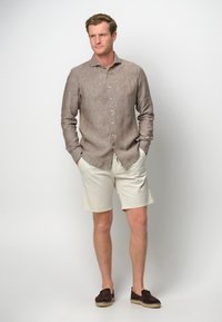 Camicia di lino marrone chiaro, pantaloni corti beige chiaro e scarpe scivolate marrone scuro; vestibilità rilassata, colletto e bottoni sulla camicia; texture semplici.