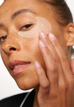 Gel eye patch being applied under the eye. Subject has freckled skin, natural makeup, and uses a neutral background. Hand gently holds the patch.