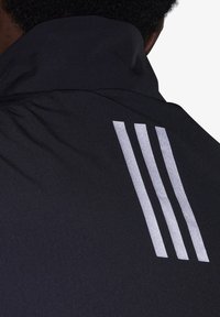 Chaqueta negra con un cuello alto y tres rayas plateadas verticales en la parte trasera. Tela suave con un ligero brillo.