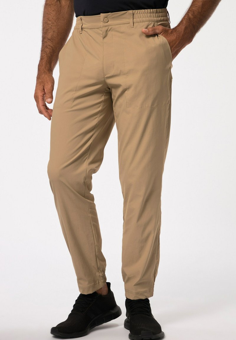 Pantalones beige con cintura elástica, dos bolsillos frontales y dobladillos en los puños. Combinados con zapatillas deportivas negras sobre un fondo neutro.