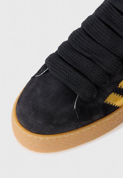 Black suede sneaker with braided laces, textured upper, yellow striped accents, and a gum rubber sole.