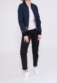 Veste aviateur navy avec des poignets et un ourlet côtelés, poche zippée sur la manche, étiquette orange sur le côté, associée à un jean noir et des baskets blanches.