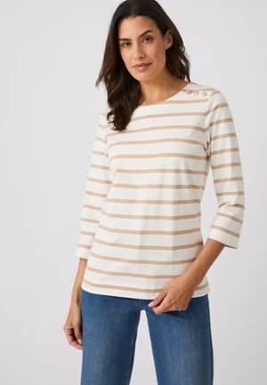 Donna che indossa una maglietta a maniche lunghe beige e bianca a righe con bottoni sulla spalla e jeans blu, in piedi davanti a uno sfondo semplice.