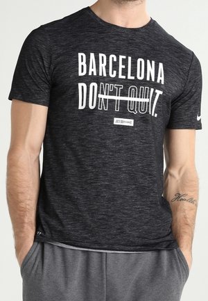 Mężczyzna w ciemnoszarej koszulce z napisem "Barcelona Don't Quit" i casualowych szarych spodniach, ręce w kieszeniach, widoczny tatuaż na przedramieniu prawej ręki.