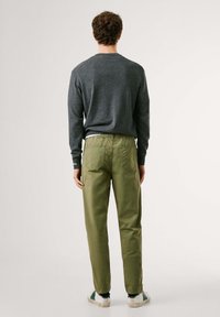 Un pull gris à manches longues, associé à un chino vert avec une ceinture élastique et deux poches arrières. Des baskets blanches et vertes sont visibles.