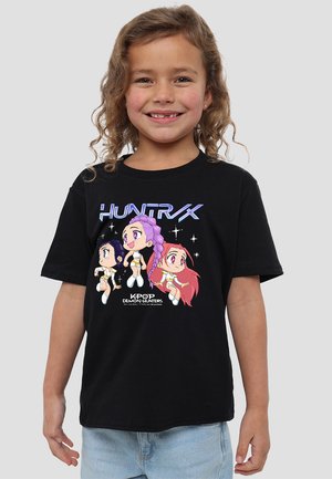 Schwarzes Baumwoll-T-Shirt mit cartoonhaften Charakteren mit bunten Haaren und Outfits. Der Druck auf der Vorderseite beinhaltet den Text "HUNTRX KPOP DEMON HUNTERS".