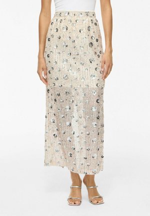 Beige midi rok met pailletten en bloemenpatroon, gedragen met een witte top en zilverkleurige sandalen met bandjes, getoond van taille tot voeten.