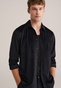 Camicia nera con bottoni, con un motivo testurizzato, maniche lunghe arrotolate, indossata sopra una maglietta nera, con chiusura a due bottoni.