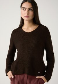 Brauner Strickpullover mit V-Ausschnitt, der diagonal gerippte Textur aufweist. Lange Ärmel und eine lockere Passform, akzentuiert durch ein kleines metallisches Logodetail.