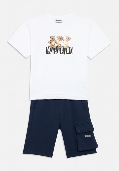 T-shirt blanc Moschino avec trois ours en peluche et logo, assorti à un short bleu marine avec une grande poche latérale arborant la marque Moschino.