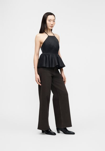 Femme debout portant un haut peplum noir à col halter, un pantalon noir à jambes larges et des bottines noires sur un fond clair uni.