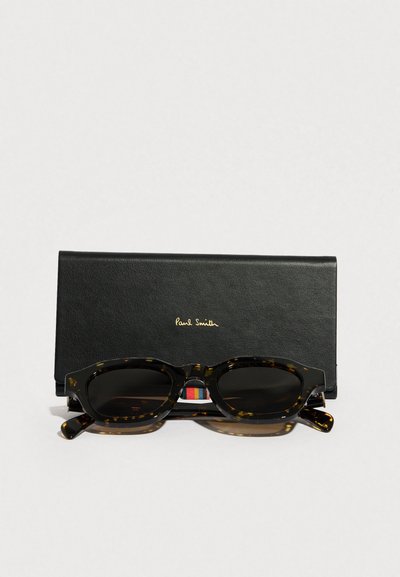 Sluneční brýle želvovinové barvy s kulatým rámem, tmavými čočkami a strukturovaným černým koženým pouzdrem s nápisem "Paul Smith" ve zlaté barvě.