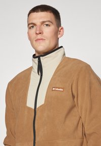 Fleecejakke i tan med en beige lynlås og høj krave. Har et kontrasterende stofindsats og et lille hvidt logo på brystet.