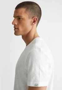T-shirt bianco con colletto rotondo realizzato in tessuto leggero. Presenta maniche corte, una leggera trama melange e un piccolo etichetta del marchio sulla manica.