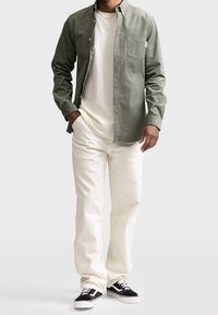 Olivgrünes Button-Up-Hemd über einem weißen T-Shirt, kombiniert mit cremefarbenen Hosen und schwarzen Sneakers mit weißen Sohlen; glatte Stofftexturen.