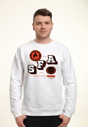 Mężczyzna ubrany w biały sweatshirt z pomarańczowymi i czarnymi logo oraz napisem "Starfleet Academy SFA", stojący na jednolitym jasnym tle.