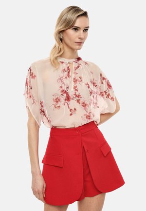 Donna con capelli biondi che indossa una camicetta floreale trasparente con scollo a goccia e pantaloncini rossi a vita alta con tasche frontali.