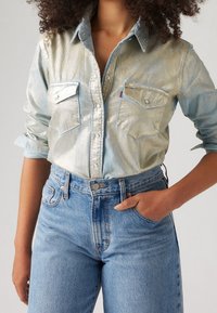 Persona in camicia metallica argento con maniche arrotolate, infilata in un paio di jeans blu a vita alta, con una mano in tasca.