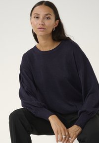 Marineblauer Pullover mit funkelndem Textil, lässiger Schnitt und weiten Ärmeln. Kombiniert mit schwarzen Hosen, inszeniert vor einem neutralen Hintergrund.