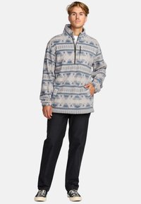 Polarowy sweter w jasnoszarym kolorze z geometrycznymi wzorami w odcieniach niebieskiego. Posiada wysoki kołnierz, zamek z przodu i kieszenie po bokach. Noszony z czarnymi spodniami.