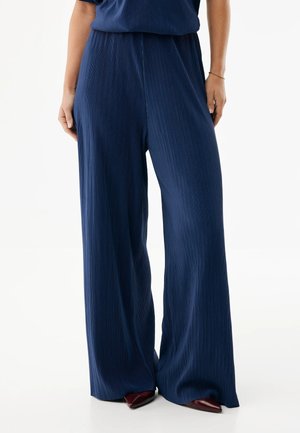 Kobieta ubrana w granatowe plisowane spodnie o szerokich nogawkach i bordowe buty w szpic, stojąca na białym tle.