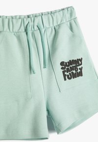 Pantaloni corti in cotone verde menta con vita elasticizzata, dotati di due tasche laterali e una grafica nera "SUNNY AND FUNNY" sul davanti.