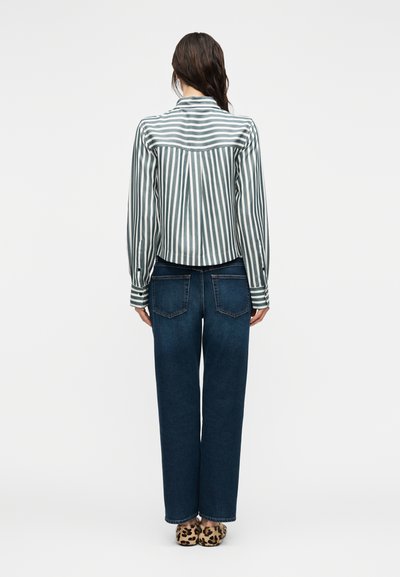 Γυναίκα που φοράει πράσινο και άσπρο ριγέ πουκάμισο, σκούρα μπλε τζιν και παντόφλες με λεοπάρ σχέδιο, στέκεται με την πλάτη στραμμένη σε λευκό φόντο.