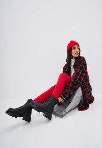 Bocanci negri până la gleznă cu un design texturat, talpă groasă, colanți roșii, jachetă din cămașă în carouri și o căciulă roșie pe un fundal alb.