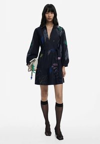 Vestido azul marino con escote en V y mangas largas abullonadas. Presenta estampados botánicos coloridos, cintura ajustada y falda de volantes. Combinado con medias negras hasta el muslo.