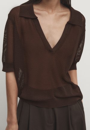 Donna che indossa un top marrone a maniche corte in maglia traforata con ampio scollo a V e colletto, abbinato a pantaloni scuri a pieghe.