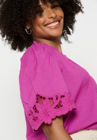 Blusa magenta con maniche corte arricciate, decorata con un intricato bordo in pizzo floreale. Il tessuto ha una texture liscia, conferendo un aspetto leggero.
