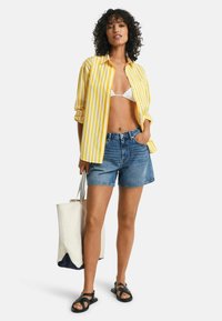 Geel en witte gestreepte shirt, witte bikini-top, denim shorts, zwarte sandalen, met een crèmekleurige en marineblauwe totebag. Krullend haar, neutrale achtergrond.