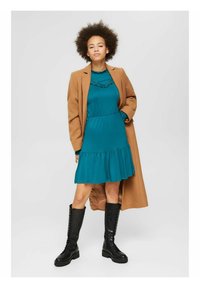 Vestido verde-água com bainha em camadas, por baixo de um longo casaco castanho. Botas pretas de atacadores e um decote redondo completam o visual. Textura suave.