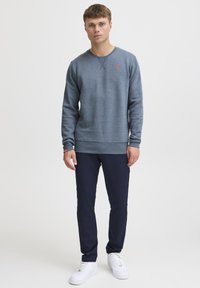 Lys grå sweatshirt med en tekstureret overflade, rund hals og orange logo, parret med navy bukser og hvide sneakers.
