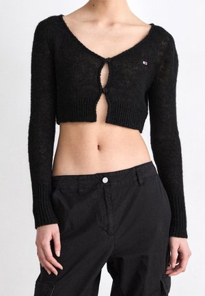 Kvinde iført en sort kort cardigan med to knapper lukket og sorte højttaljede bukser, der viser maven og arme i siderne.