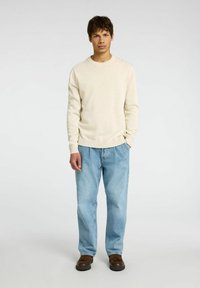 Hellbeige Crewneck-Pullover mit gerippten Bündchen und Saum, kombiniert mit lockeren hellblauen Jeans und brauen Schuhen. Minimalistisches Design.