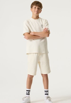 Cream-colored cotton short-sleeve shirt and matching shorts, featuring a small logo detail. Worn with white sneakers and black patterned socks.