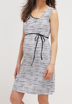 Femme enceinte portant une robe blanche sans manches, longueur genoux, avec des rayures horizontales noires et une ceinture noire nouée au-dessus du ventre.