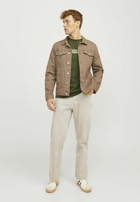 Chaqueta de mezclilla marrón sobre una camiseta verde, con jeans rectos de un color beige claro y zapatillas blancas con rayas negras y verdes.