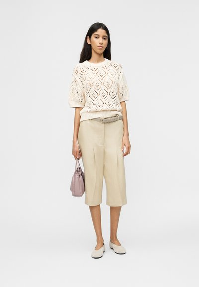 Γυναίκα με κρεμ πλεκτό πουλόβερ κοντομάνικο, μπεζ κολάν με ζώνη, κρεμ φλατ παπούτσια, κρατώντας μικρή τσάντα χειρός σε χρώμα μωβ, στέκεται μπροστά σε απλό φόντο.