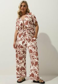 Tvådelad outfit bestående av en kortärmad skjorta med knappar och breda byxor. Tyget är mönstrat med bruna blommönster på en krämfärgad bakgrund.